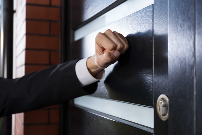 A close-up image of a hand, with a closed fist, knocking on a door.