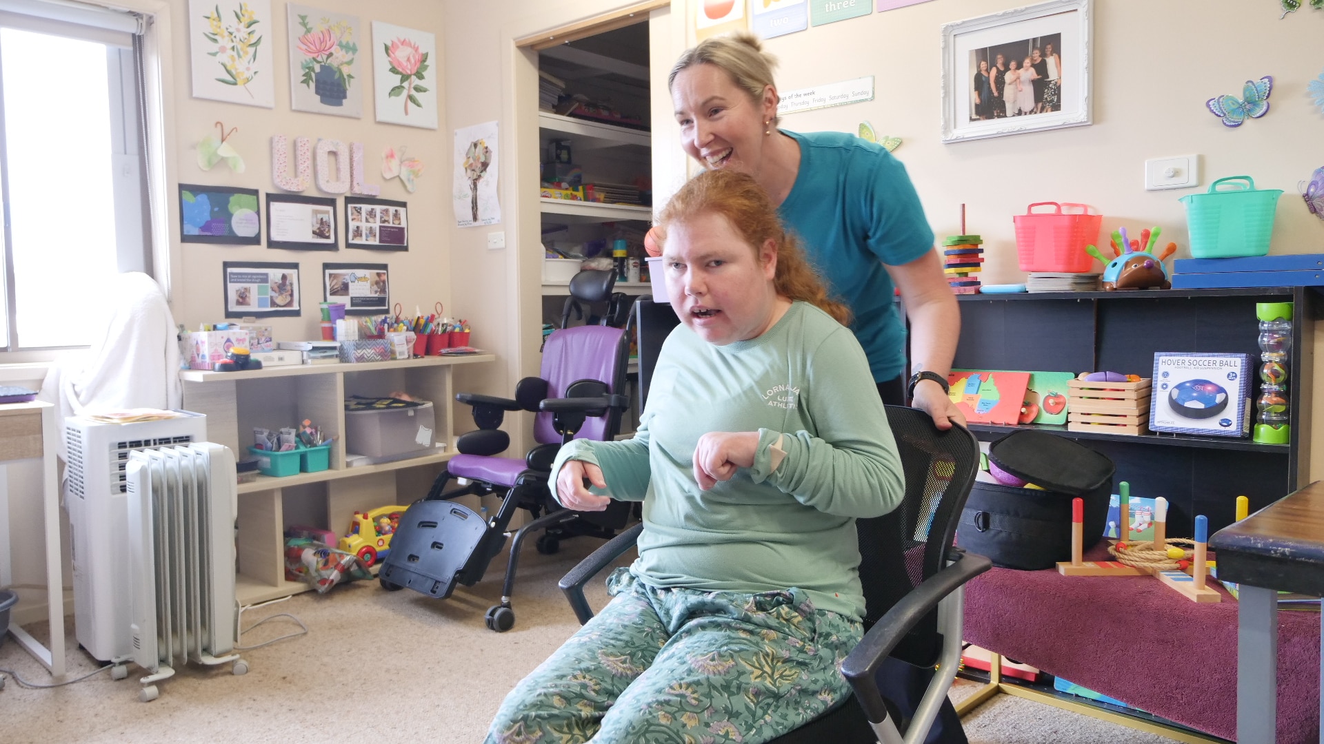 A young woman sitting in a wheelie chair being pushed by another woman inside a room with colourful objects on shelves 