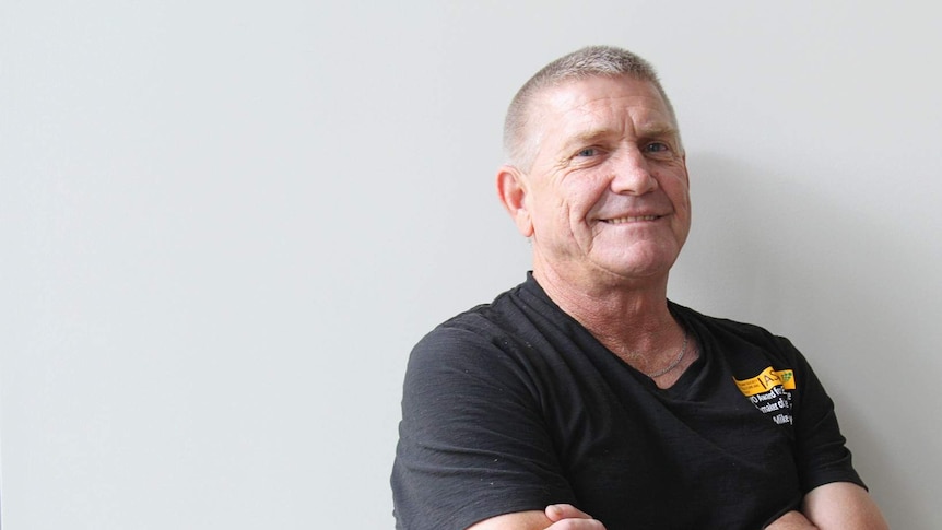 Man with grey hair in a black shirt in front of white wall.