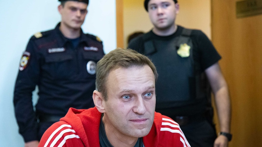 A man wearing a red jacket stares ahead with two guards behind him.