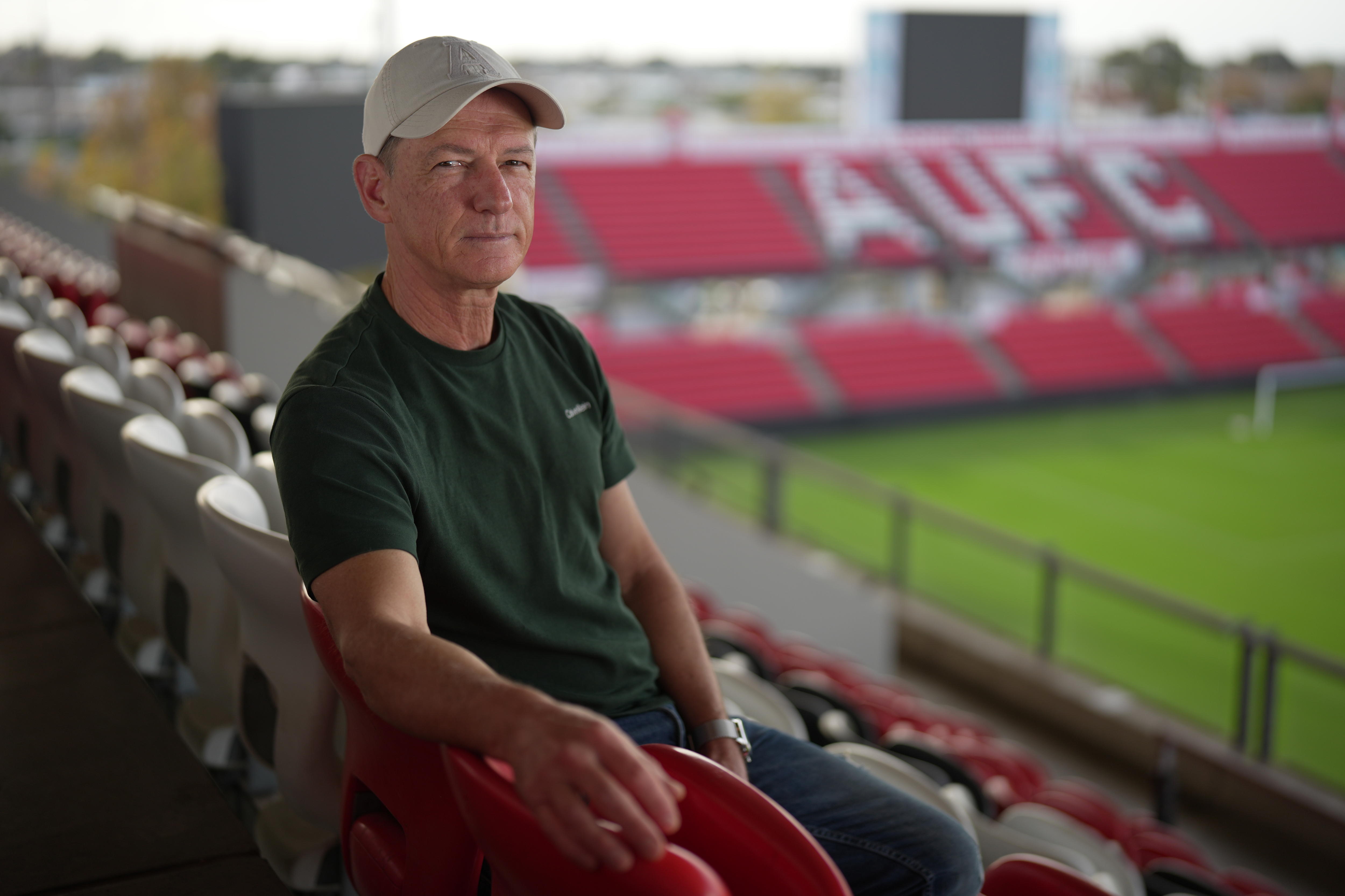 A man in a cap sits in the audience seats overlooking a soccer field with AUFC emblazoned in the audience stand