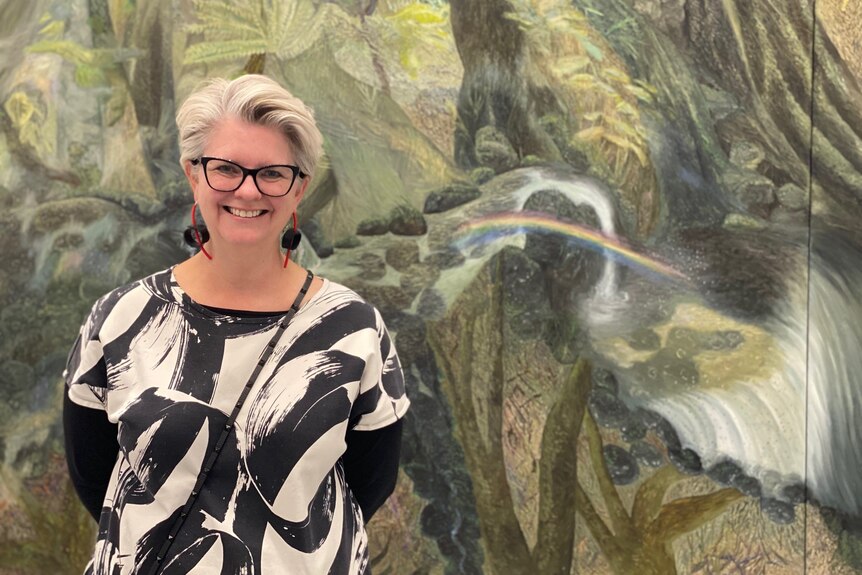 Woman wearing glasses smiling in front of a wall-length painting of a rainforest with a rainbow through waterfalls
