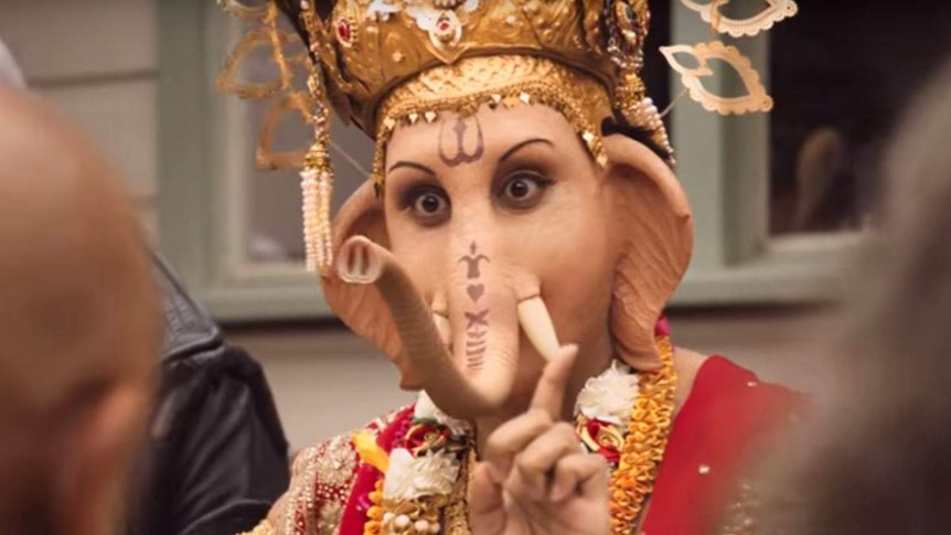 A man dressed as the God Ganesha holds up his finger. He has the head of an elephant.