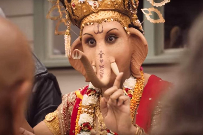 A man dressed as the God Ganesha holds up his finger. He has the head of an elephant.