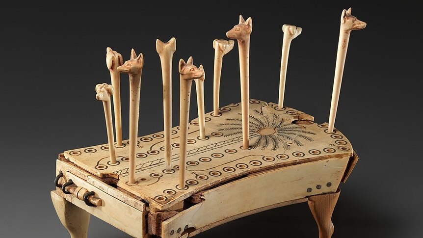 A light timber platform with a pattern of multiple holes on top. There are long sticks with animal heads carved into them.