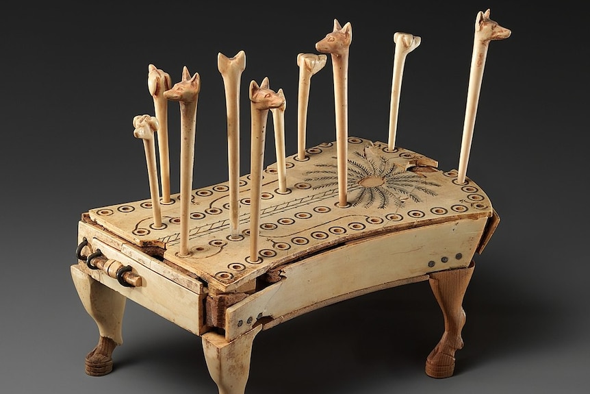 A light timber platform with a pattern of multiple holes on top. There are long sticks with animal heads carved into them.