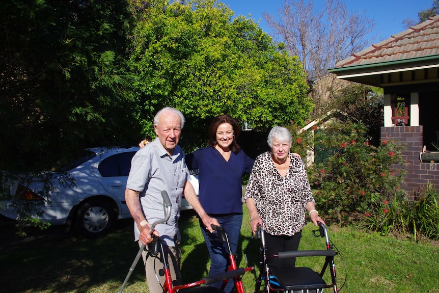 Christine Hall stands with her elderly parents on either side of her, the a garden outside their home