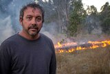 An Indigenous man stands in front of a control-burned grassfire