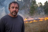 An Indigenous man stands in front of a control-burned grassfire