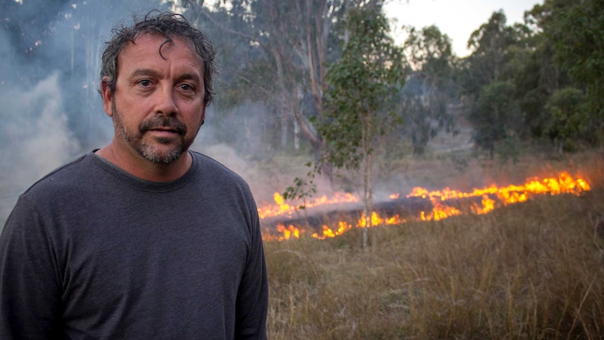 An indigenous man stands in front of a fire burning in the bush.