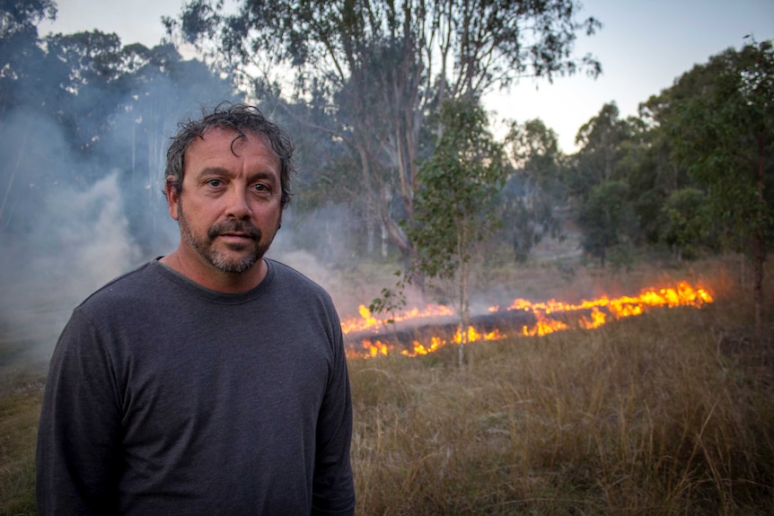 An Indigenous man stands in front of a control-burned grassfire