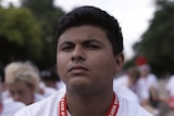 A male teen with serious expression sits outdoors on ground near trees, among many boys in white t-shirts with red lanyards.