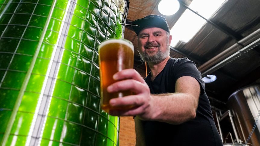 a man smiling and holding a beer next to a large vat with glowing green liquid