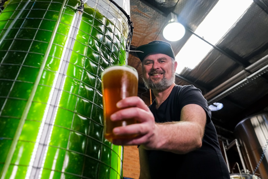 a man smiling and holding a beer next to a large vat with glowing green liquid