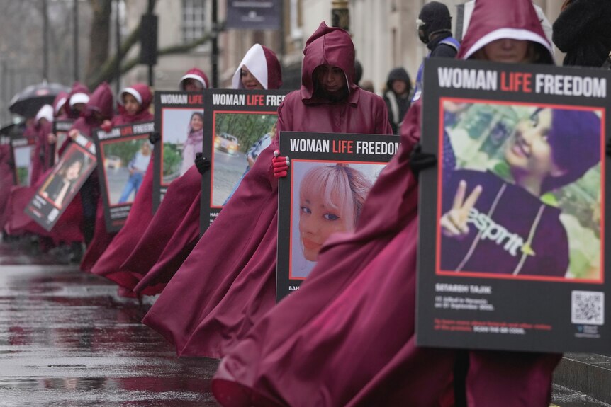 Women wearing maroon Handmaid's Tale cloaks and white wimples march, holding placards aloft.