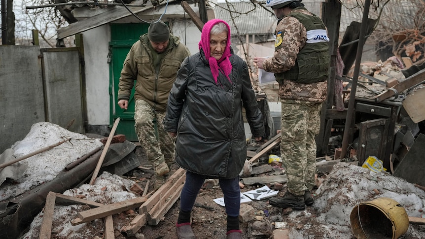 An elderly woman walks thorugh the ruins of her home.