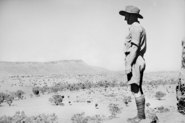 A black-and-white image of a soldier standing on a small hill and looks over a vast Central Australian landscape.