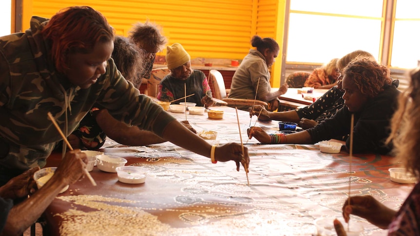 Aboriginal women paint around a canvas in a corrugated building in a remote community.