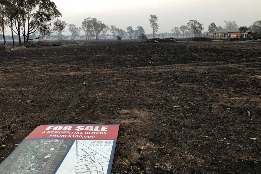 A residential for sale sign lies flat on the burnt ground with smoke in the air.