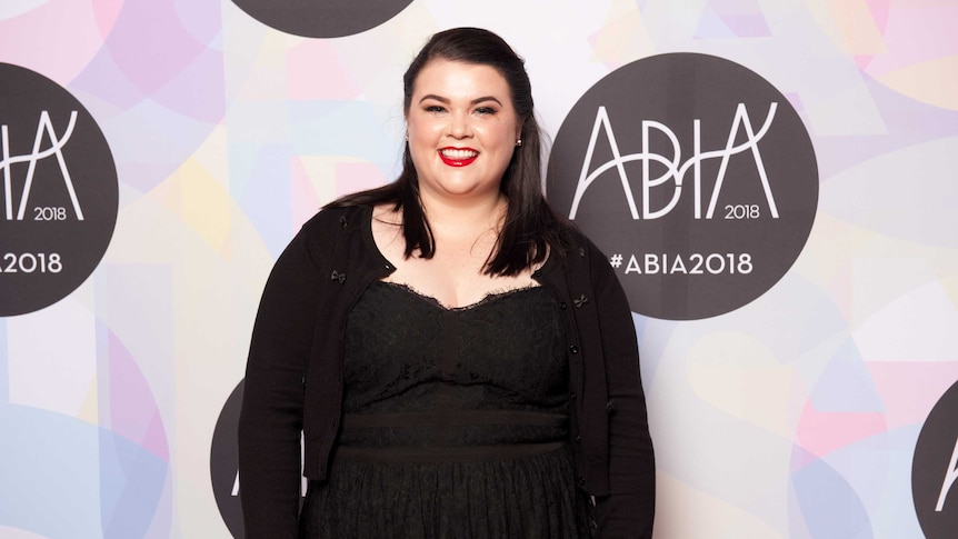 The author, wearing a black dress and bright red lipstick, stands against a pastel coloured media wall.