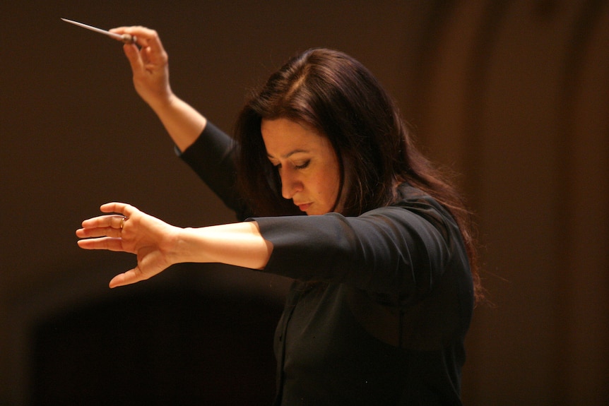 White middle-aged woman with long brown hair wears all black and stands on a podium wielding a conductor's baton.