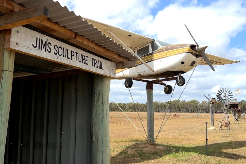 A woodern hut with Jims Scultpure Trail sign. A plane on a pole is in the background, with a windmill further back.
