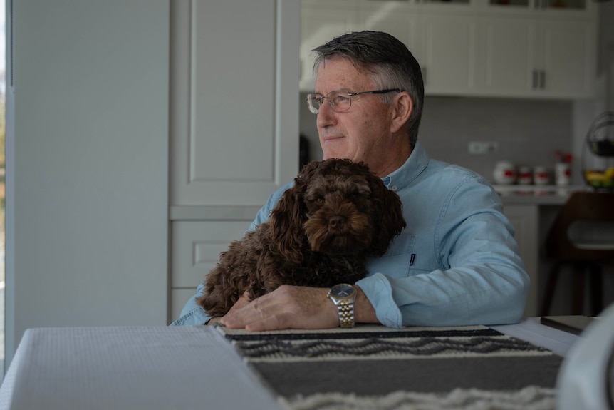 An older man peers out his window. His chocolate brown cavoodle is on his lap and looks directly at the camera