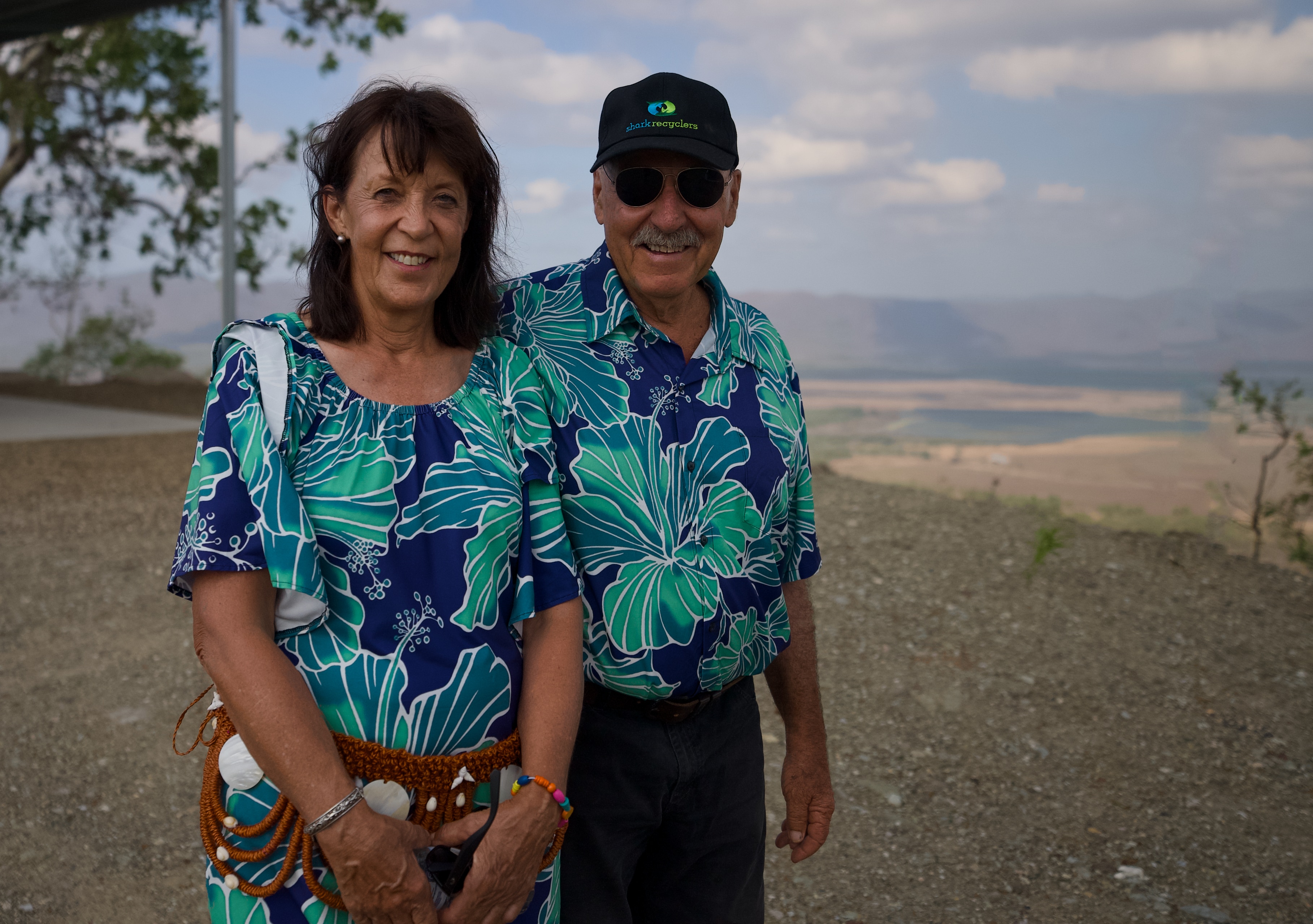 Una pareja sonriente se encuentra afuera, en una colina con tierras de cultivo que se extienden a lo lejos.