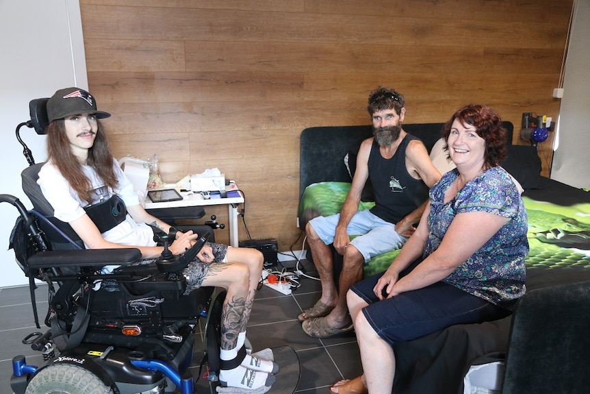 A young man in a wheelchair sitting next to a man and a woman sitting on a bed.