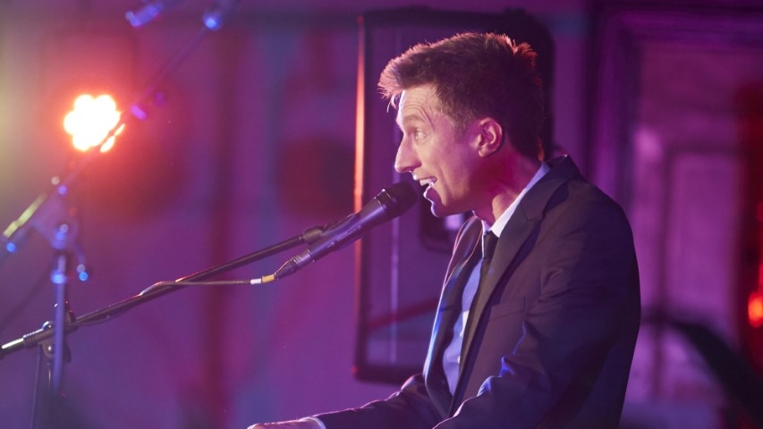 Sammy J, wearing a suit and tie, sings at the piano