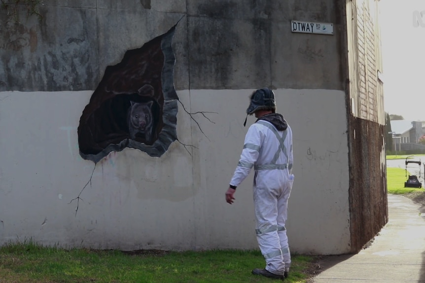 A strangely dressed man looks at a drawing of a wombat emerging from a cave drawn on a bridge