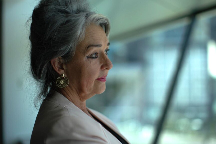 A woman with grey hair looks out a window at a street and buildings. She has a neutral expression.