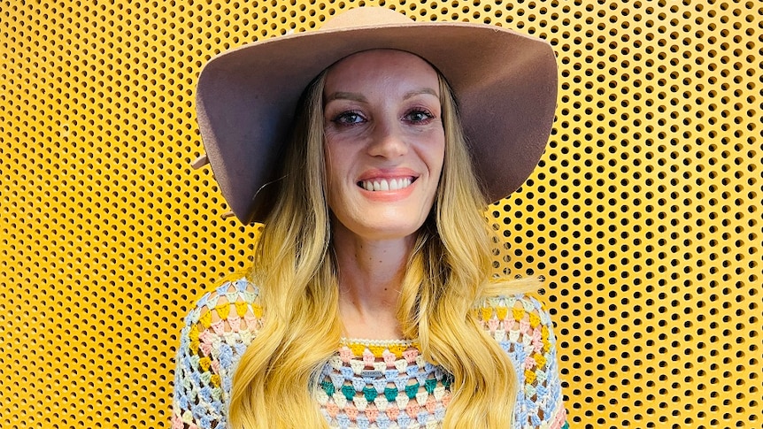 A woman wearing a floppy hat and colourful crochet jumper smiles and looks at the camera