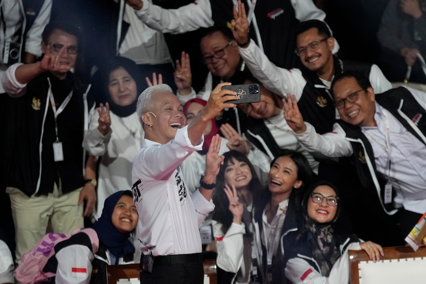 A presidential candidate holds up his phone posing for a selfie with a crowd of supporters. 