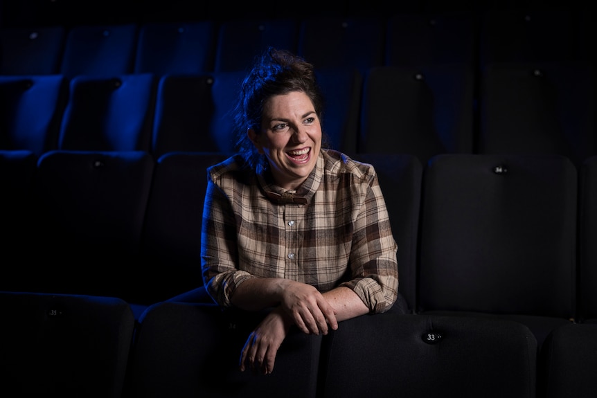 A brown-haired woman in her late 30s sits in a row of empty theatre chairs. She is smiling brightly. 