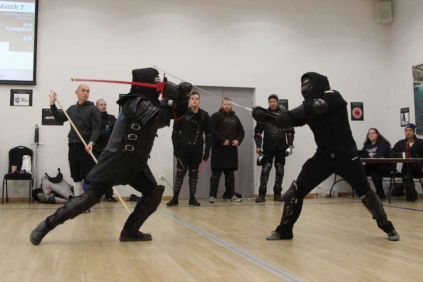 Two men dressed in black protective gear engage in sword fight while a small crowd looks on.