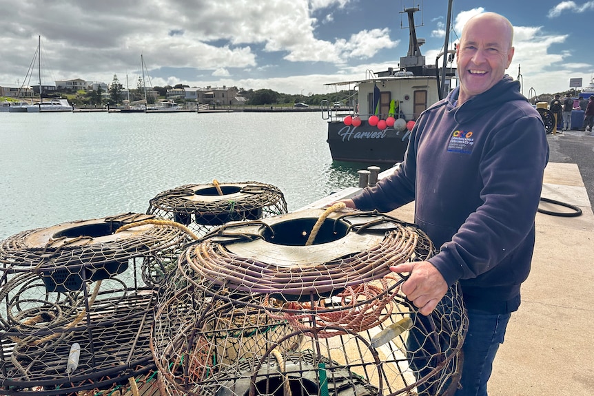 A smiling man in a hoodie stands holding a lobster pot on the edge of a wharf.