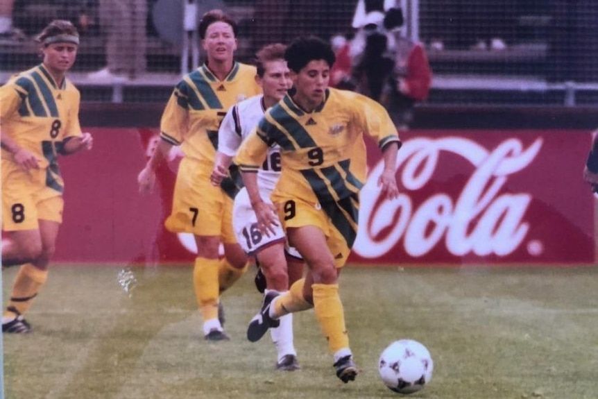 A soccer player wearing yellow and green runs with a ball during a big match with team-mates behind her
