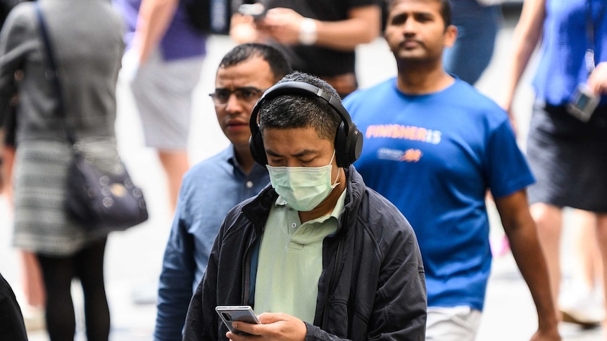 A man is wearing a protective face mask and walking while looking at his phone, he also has headphone on