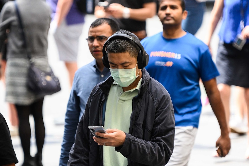 A man is wearing a protective face mask and walking while looking at his phone, he also has headphone on