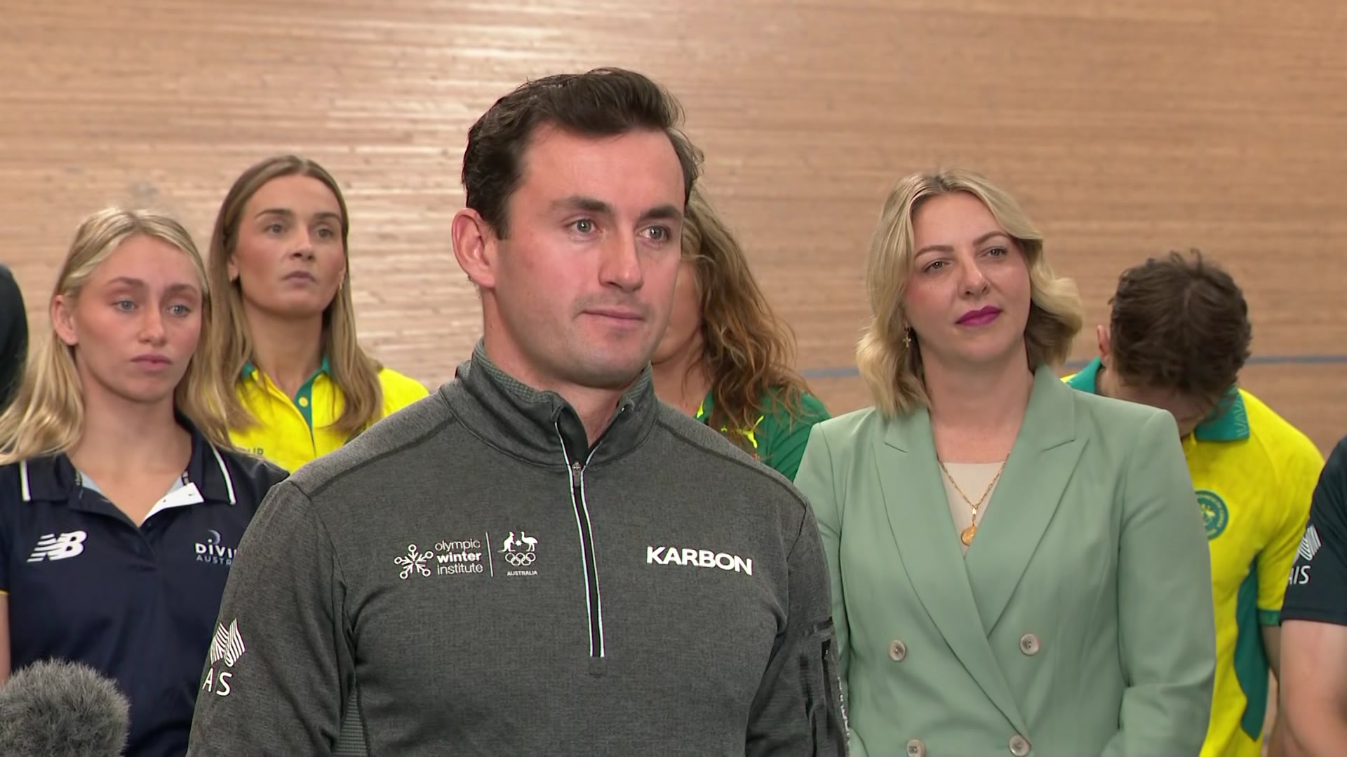 A man in a tracksuit standing in front of wooden wall with women behind him.