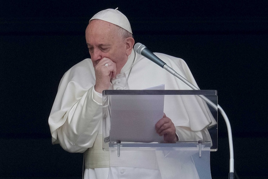 A man wearing a white frock and white skull cap, puts his hand to his mouth and coughs at a podium.