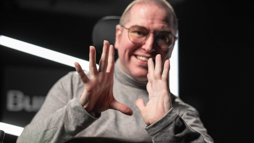 A man in a wheelchair wearing a grey turtleneck hold up his hands for the camera