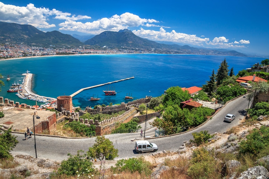 A road wraps around a bright blue harbour with mountains in the background