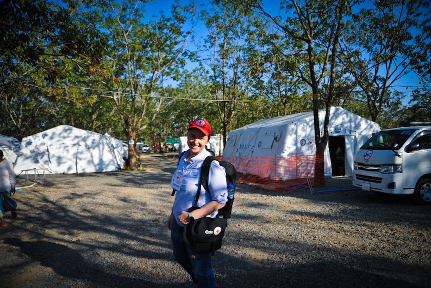 A woman with a red cross hat on stands in front of a truck and some large tents