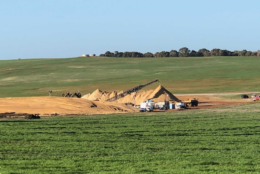 A quarry with piles of yellow sand sits in the middle of a lush green paddock on a cloudless day.