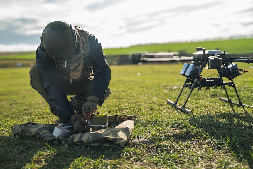 A man wearing a military uniform crouches down near a drone.