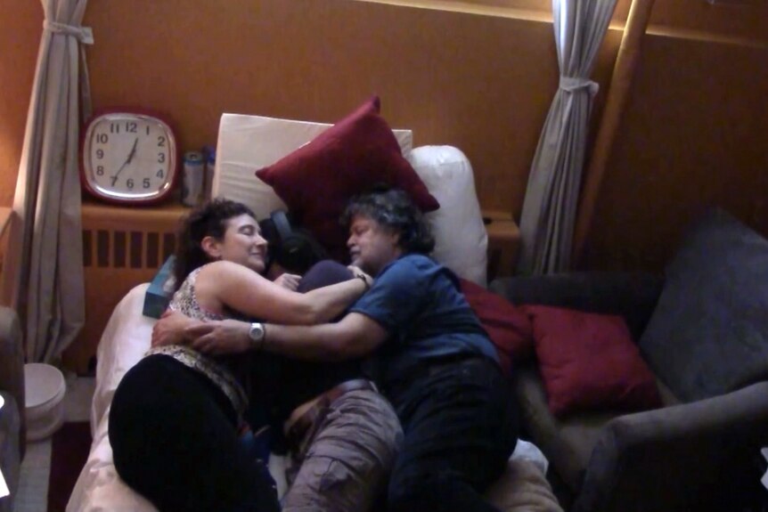 Meaghan Buisson lies on a bed with a man and woman lying on either side cuddling her. 