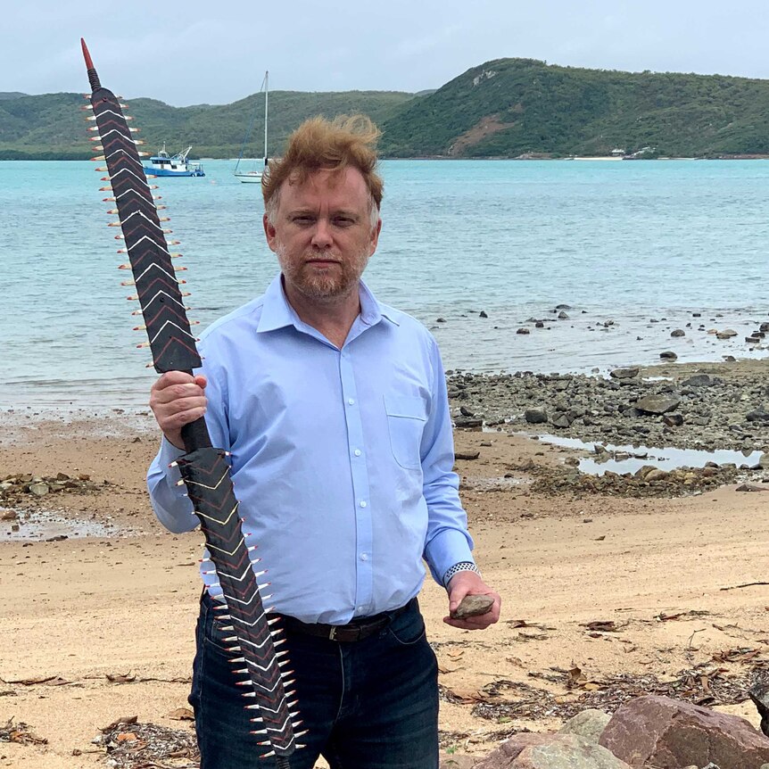 A man holds a traditional shark-tooth studded weapon. Behind him is an island with a line of red soil down its hillside.