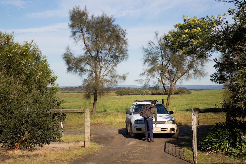 A student stands in front of his parent's car in a dirt driveway, bush and pasture surrounding.